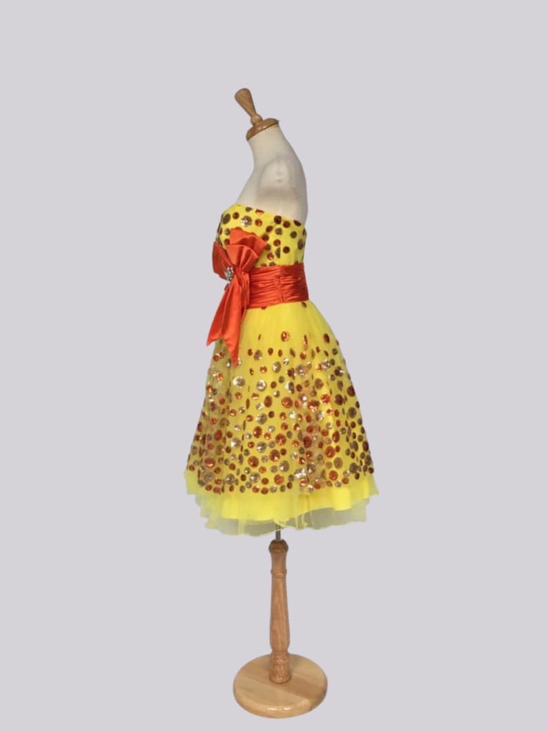 May include: A yellow tulle dress with a red satin bow and gold and orange sequins. The dress is on a wooden mannequin.