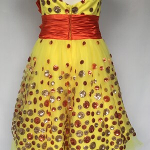May include: A yellow tulle dress with a strapless neckline and a red satin sash. The dress is covered in gold and orange sequins in a polka dot pattern.