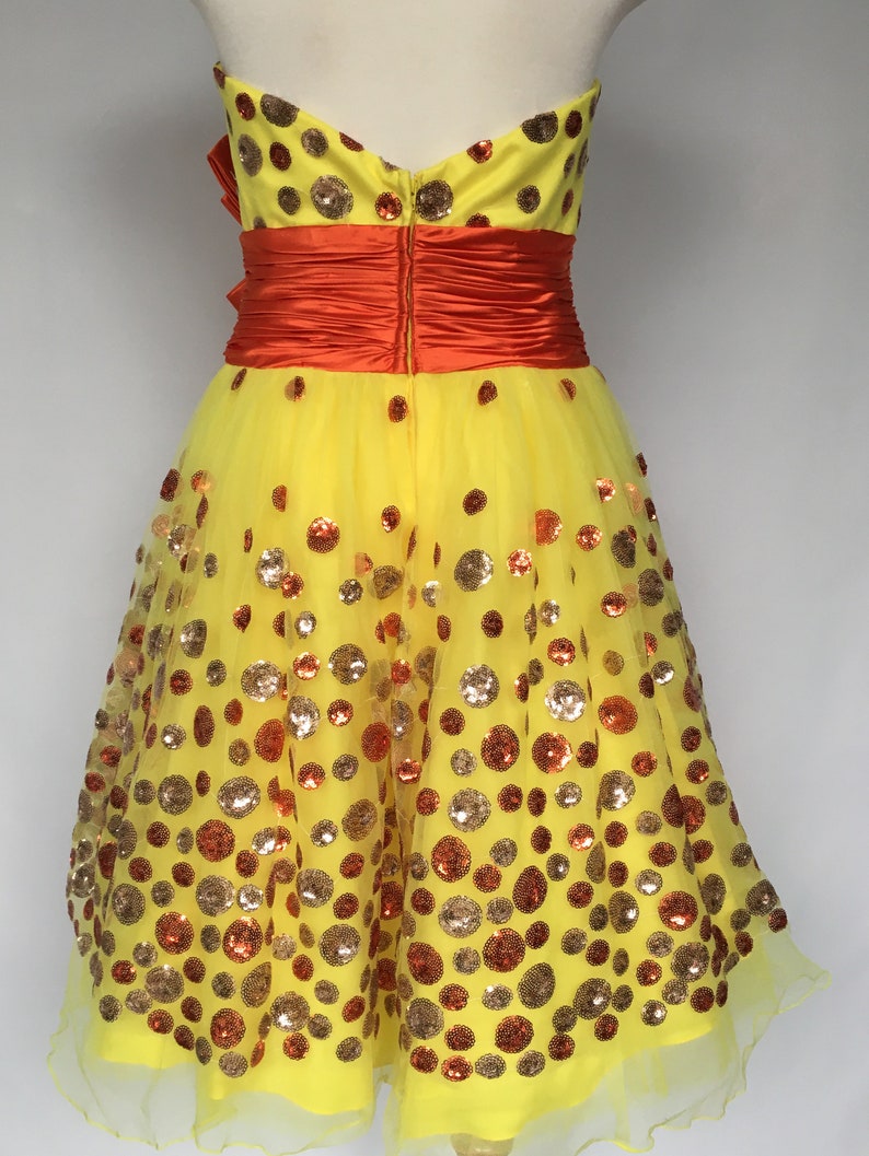 May include: A yellow tulle dress with a strapless neckline and a red satin sash. The dress is covered in gold and orange sequins in a polka dot pattern.