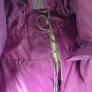 May include: Close-up of a shiny, purple garment with a drawstring closure. The fabric has a glossy appearance, with visible stitching and a zipper detail. The garment appears to be a jacket or coat, with a light-colored lining visible.