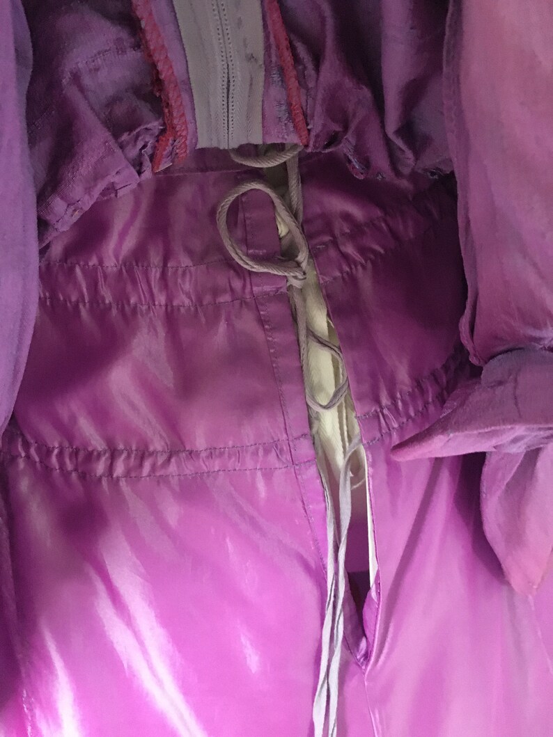 May include: Close-up of a shiny, purple garment with a drawstring closure. The fabric has a glossy appearance, with visible stitching and a zipper detail. The garment appears to be a jacket or coat, with a light-colored lining visible.