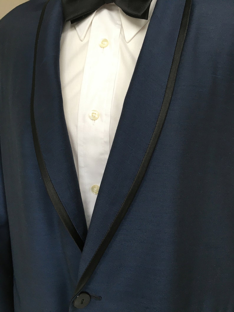 May include: A dark blue tuxedo jacket with a black satin lapel. The jacket has a single button closure and a white dress shirt underneath.