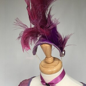 May include: A purple and pink feather headdress with a matching choker necklace. The headdress features a curved band adorned with feathers in shades of purple and pink. The choker has a purple pendant.