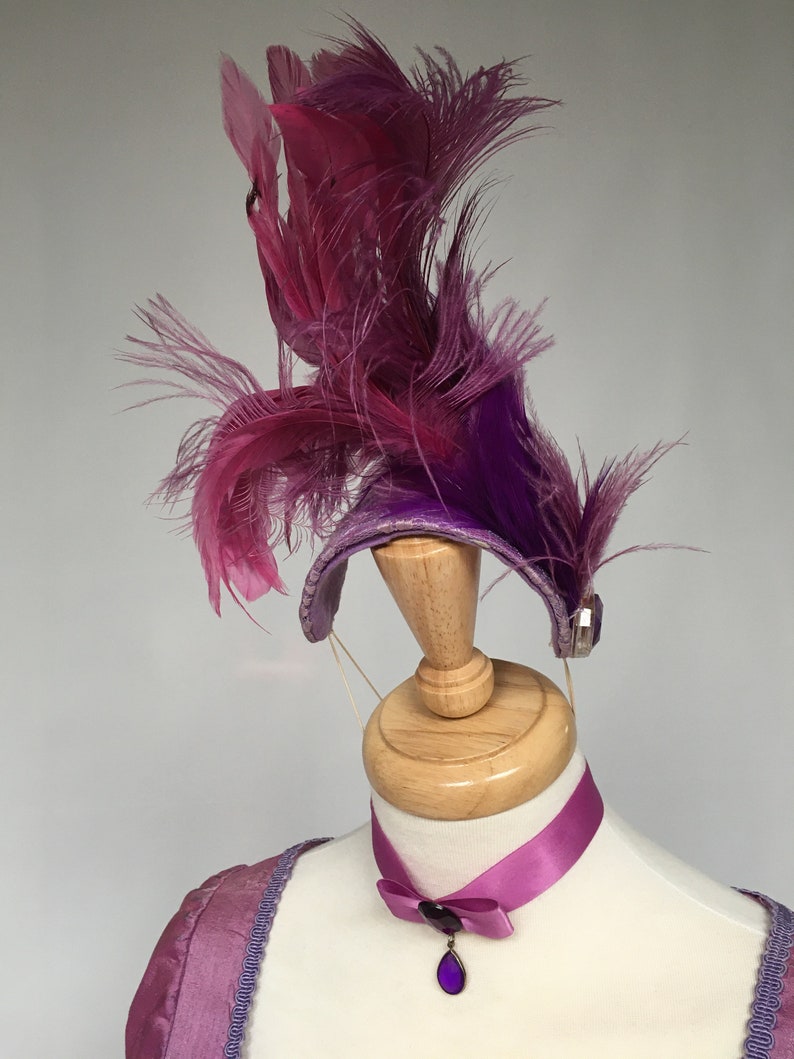 May include: A purple and pink feather headdress with a matching choker necklace. The headdress features a curved band adorned with feathers in shades of purple and pink. The choker has a purple pendant.