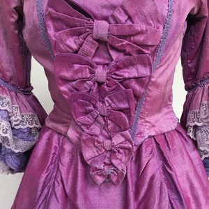 May include: A vintage-style, magenta-colored dress with a square neckline and three large bows cascading down the bodice. The dress features ruffled sleeves with lace trim and a textured fabric, evoking a historical aesthetic.
