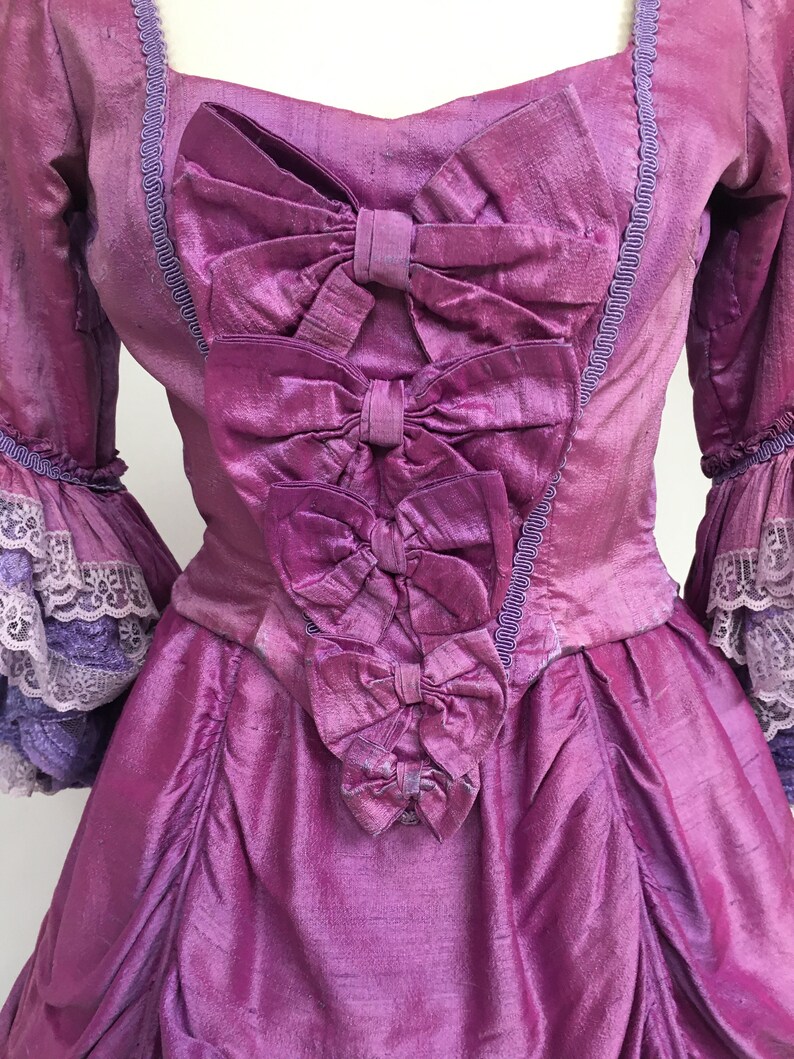 May include: A vintage-style, magenta-colored dress with a square neckline and three large bows cascading down the bodice. The dress features ruffled sleeves with lace trim and a textured fabric, evoking a historical aesthetic.