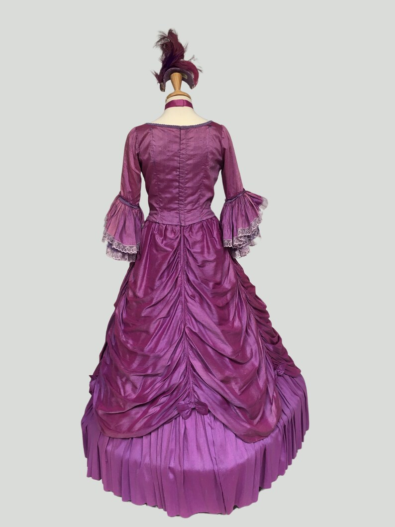 May include: A vintage purple gown with a matching feathered headpiece. The dress features long sleeves with ruffled cuffs, a fitted bodice, and a full skirt with intricate draping. The gown is displayed on a mannequin, showcasing its historical design.