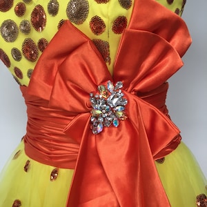 May include: A yellow tulle dress with orange satin ribbon and a large jeweled bow. The dress is covered in small brown and orange sequins.