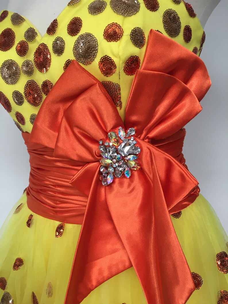 May include: A yellow tulle dress with orange satin ribbon and a large jeweled bow. The dress is covered in small brown and orange sequins.