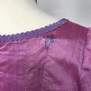 May include: Close-up of a magenta-colored dress bodice with a textured, iridescent fabric. The neckline is trimmed with a decorative, wavy, purple trim. A matching purple ribbon is visible at the neck.