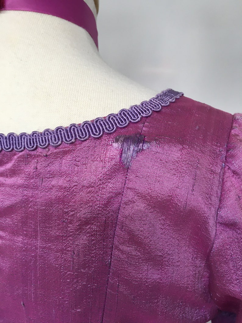 May include: Close-up of a magenta-colored dress bodice with a textured, iridescent fabric. The neckline is trimmed with a decorative, wavy, purple trim. A matching purple ribbon is visible at the neck.