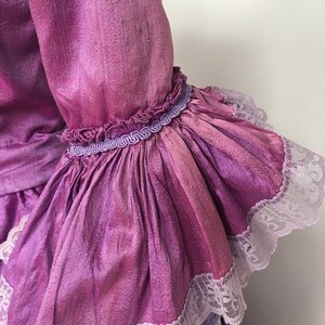 May include: Close-up of a vibrant, iridescent purple sleeve with ruffled cuffs. The sleeve is adorned with delicate white lace trim and a decorative purple trim. The fabric has a textured appearance, suggesting a silk or satin material.