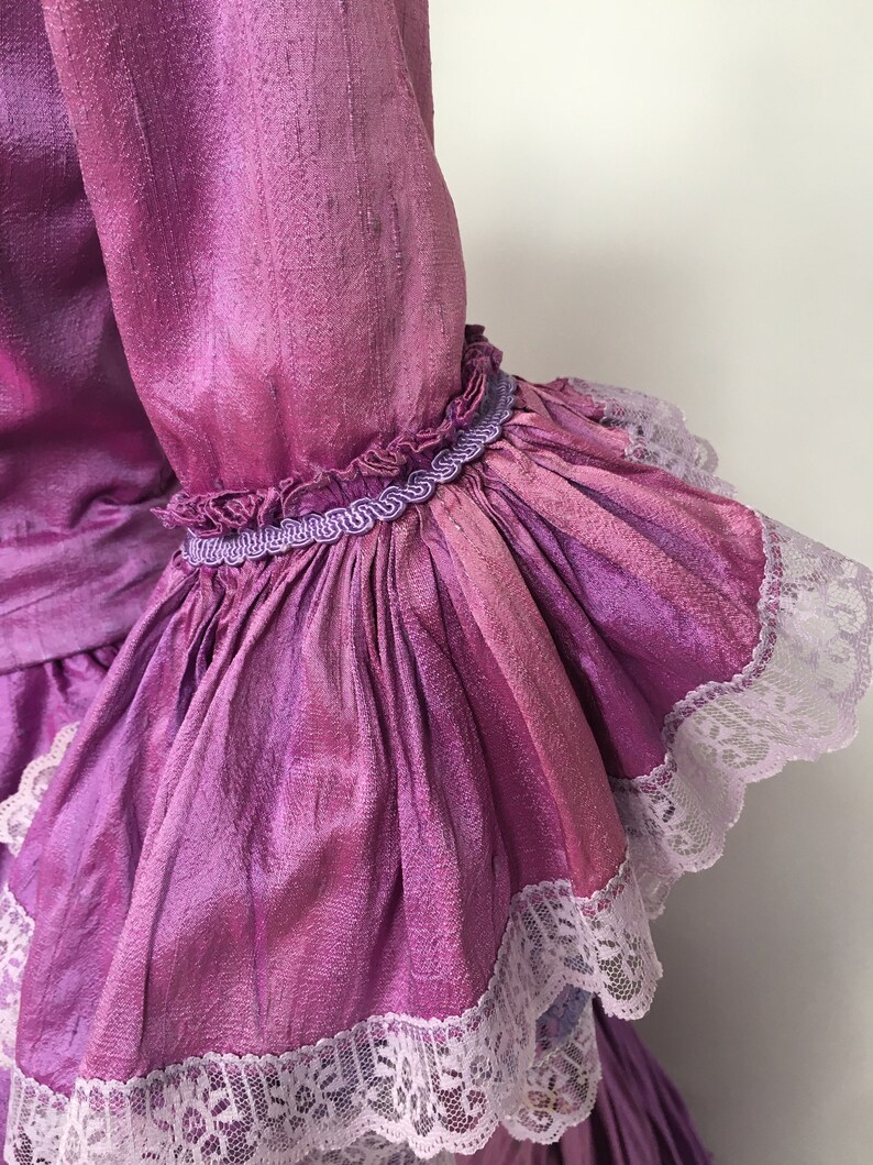 May include: Close-up of a vibrant, iridescent purple sleeve with ruffled cuffs. The sleeve is adorned with delicate white lace trim and a decorative purple trim. The fabric has a textured appearance, suggesting a silk or satin material.