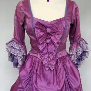May include: A vibrant purple historical gown with a matching choker. The dress features a square neckline, bows, and ruffled sleeves with lace trim. The fabric has a shimmering texture, and the choker has a pendant.
