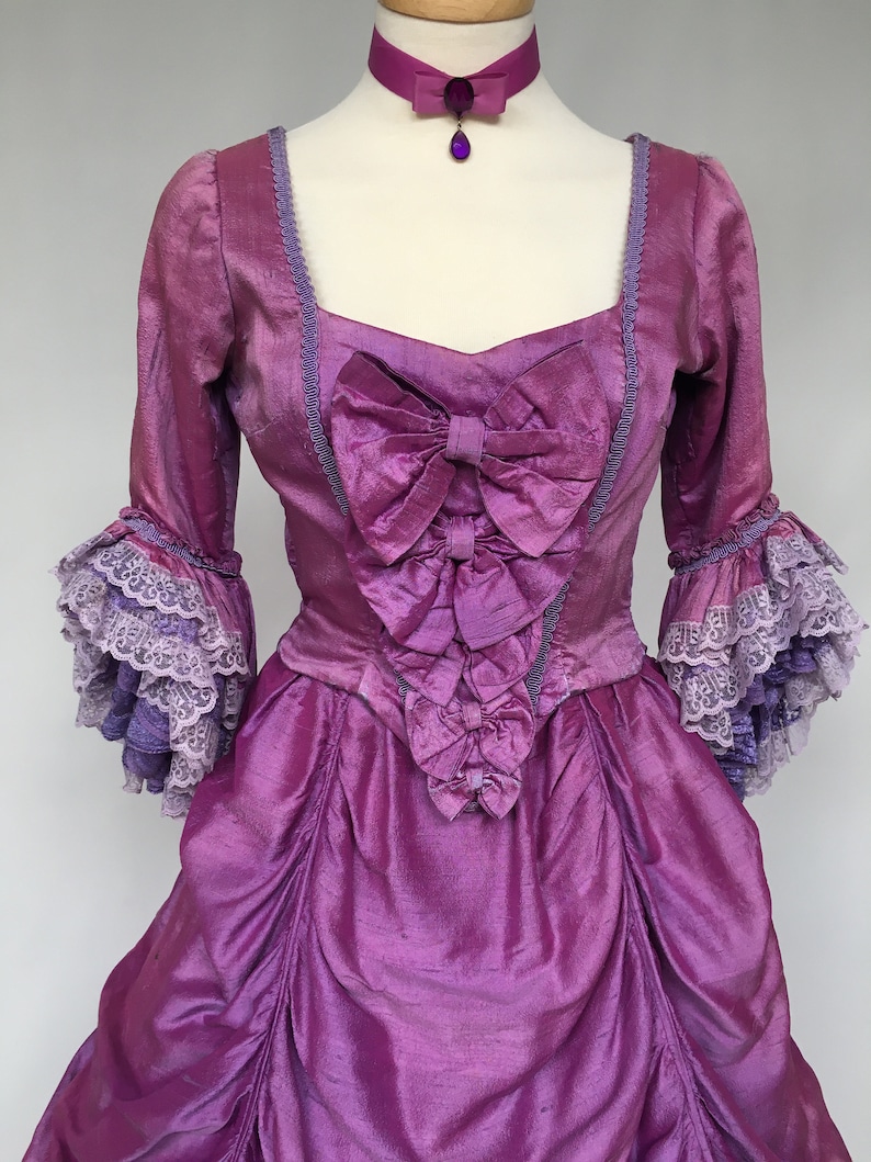 May include: A vibrant purple historical gown with a matching choker. The dress features a square neckline, bows, and ruffled sleeves with lace trim. The fabric has a shimmering texture, and the choker has a pendant.