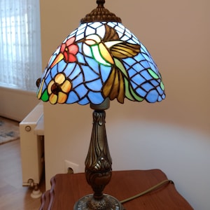 May include: A stained glass table lamp with a blue, green, and yellow hummingbird design. The lamp has a bronze base with intricate details.