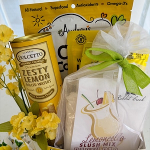 May include: A gift basket filled with lemon-themed treats. It includes a can of Dolcetto Zesty Lemon rolled wafers, a bag of lemon-flavored mix, and other packaged snacks. The basket is decorated with yellow flowers and a green bow. The text "Easy Peasy Lemon" is visible.