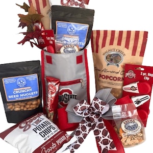 May include: A gift basket filled with snacks and treats, including bags of potato chips, popcorn, and crunchy beer nuggets. The basket features a gray and red Ohio State-themed cooler bag, a decorative bow, and various other goodies.