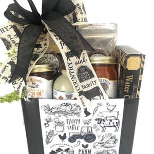 May include: A gift basket with a black and white farm theme. The basket contains jars of jam and other food items, tied with a black ribbon. The basket is decorated with illustrations of farm animals and the words "Farm to Table".