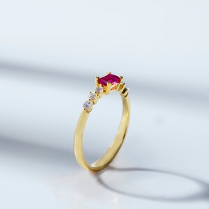 May include: A gold ring with a red gemstone and small clear gemstones. The ring is set on a white background.