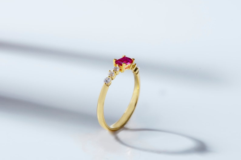May include: A gold ring with a red gemstone and small clear gemstones. The ring is set on a white background.
