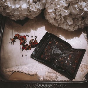 May include: A black pouch filled with loose leaf tea, labeled "Blood Moon Black Tea" with ingredients listed as "Black Tea, Hibiscus, Rose Petals, Lavender, Rose Buds". The pouch is on a white tray with scattered red rose petals.
