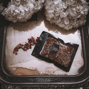 May include: A black pouch filled with dried herbs and flowers, labeled "Love Potion" with a floral design. The pouch is on a dark, ornate tray with white hydrangeas in the background.