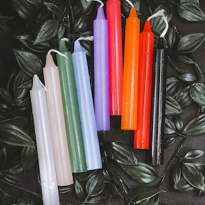 Könnte beinhalten: Ein Set aus acht dünnen, spitz zulaufenden Kerzen in verschiedenen Farben, darunter Weiß, Grün, Blau, Lila, Rosa, Orange, Rot und Schwarz. Die Kerzen sind in einem Halbkreis auf einem dunklen Hintergrund mit grünen Blättern angeordnet.