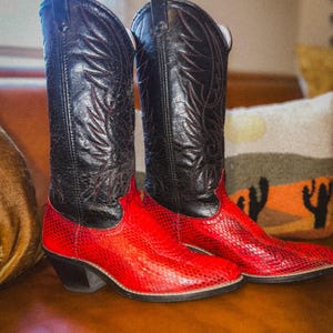 May include: A pair of cowboy boots with a striking color contrast. The boots feature black leather uppers with decorative stitching and red, textured lower sections. The boots have a classic western design with angled heels.