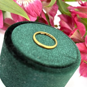 Puede incluir: Un anillo de oro descansa sobre una superficie de terciopelo verde, con flores rosas y blancas en el fondo. El anillo es de diseño de banda simple, con marcas visibles. Una pieza de joyería clásica.