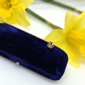 Puede incluir: Un prendedor de sombrero dorado con una cabeza en forma de flor, con un diamante central. El prendedor se exhibe en una caja de joyería de terciopelo azul oscuro, con narcisos amarillos en el fondo.