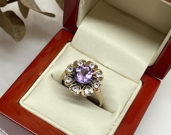 May include: A gold ring with a purple amethyst gemstone surrounded by smaller white gemstones in a flower design. The ring is in a red velvet ring box.