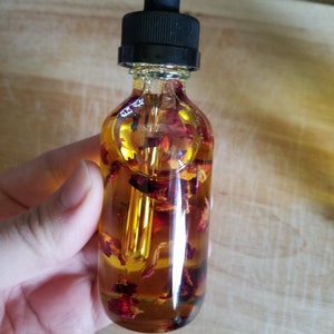 Puede incluir: Una botella de vidrio transparente con un gotero negro en la parte superior llena de un l&iacute;quido amarillo y p&eacute;talos de rosa rojos.