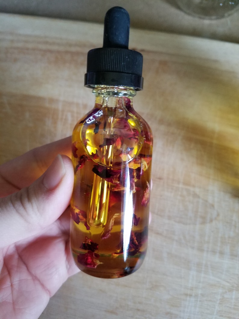 Puede incluir: Una botella de vidrio transparente con un gotero negro en la parte superior llena de un l&iacute;quido amarillo y p&eacute;talos de rosa rojos.