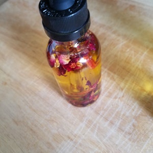 Puede incluir: Una botella de vidrio transparente con un gotero negro en la parte superior llena de un l&iacute;quido amarillo y p&eacute;talos de rosa secos.