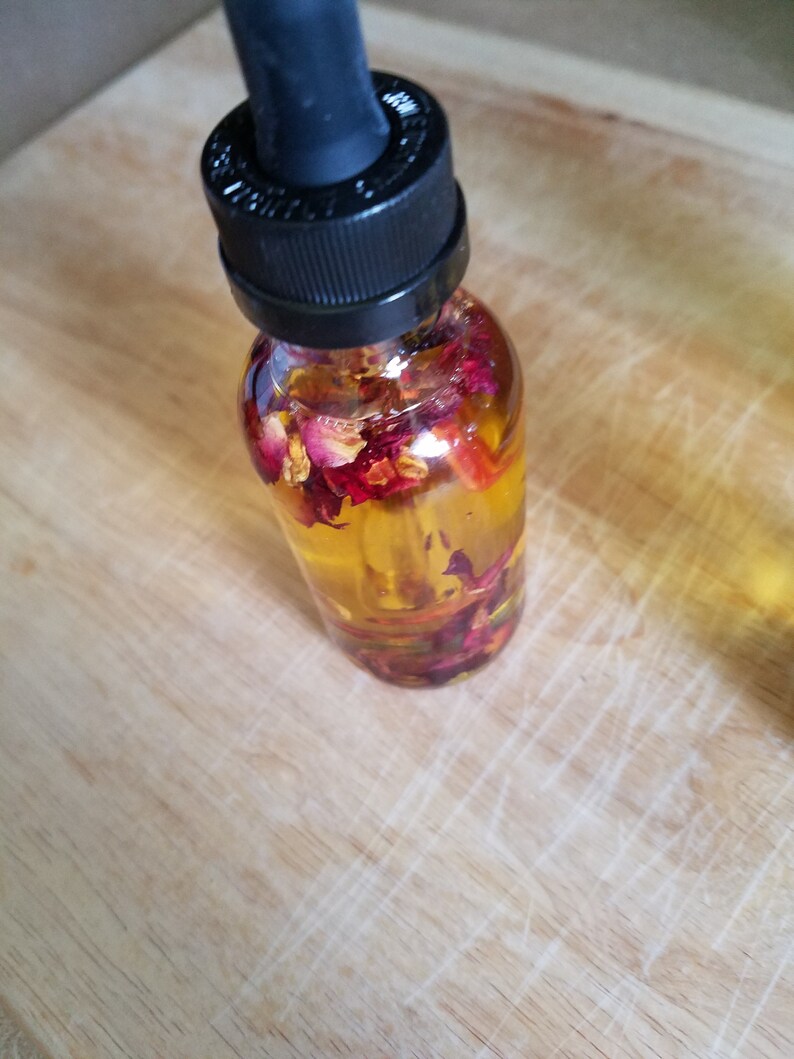Puede incluir: Una botella de vidrio transparente con un gotero negro en la parte superior llena de un l&iacute;quido amarillo y p&eacute;talos de rosa secos.
