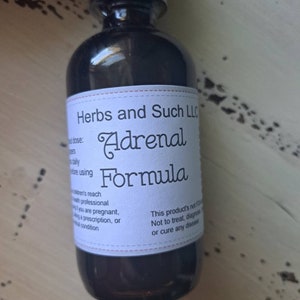 May include: A dark brown glass bottle with a black label that reads "Herbs and Such LLC Adrenal Formula". The label also includes a disclaimer that the product is not FDA approved and does not treat, diagnose, or cure any disease.