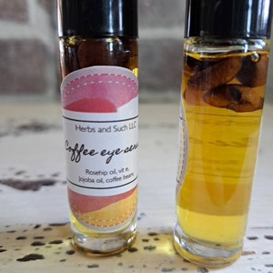 May include: Two clear glass bottles with roll-on tops filled with a light brown liquid. The bottles have labels with the text "Herbs and Such LLC" and "Coffee eye serum". The label also lists the ingredients: "Rosehip oil, vit e, Jojoba oil, coffee beans".