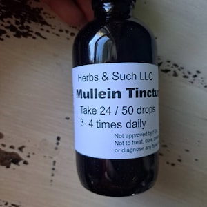 May include: A dark glass bottle of Mullein Tincture with a black cap. The white label reads "Herbs & Such LLC" and dosage instructions. The bottle is held by a person.