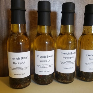 May include: Four glass bottles of French Bread Dipping Oil with black lids. The labels are white with black text and list the ingredients: olive oil, garlic, kosher salt, red pepper, dill weed, parsley, basil, oregano. The bottles are filled with a clear, golden oil and contain herbs and spices.