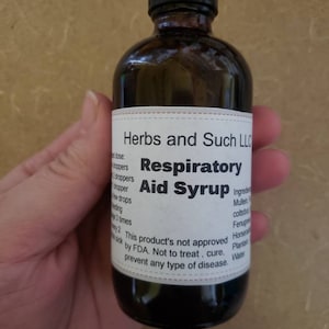 May include: A dark brown glass bottle with a dropper lid. The label reads "Herbs and Such LLC Respiratory Aid Syrup" and lists ingredients including Mullein, Coltsfoot, Fenugreek, Horsetail, Plantain, and Water. The label also states "This product's not approved by FDA. Not to treat, cure, prevent any type of disease."