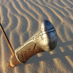 May include: Close-up of a silver metal sword hilt with intricate carvings and a textured surface. The hilt is partially buried in sand.