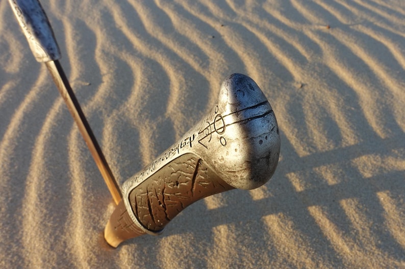 May include: Close-up of a silver metal sword hilt with intricate carvings and a textured surface. The hilt is partially buried in sand.