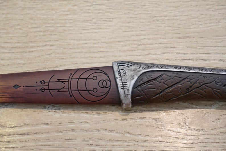 May include: A wooden and metal dagger with a decorative design featuring a circle with smaller circles inside. The handle is brown wood with a dark brown stain and the blade is silver with a textured surface.