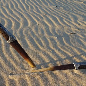 May include: Two wooden-handled knives with silver accents are lying on a sandy surface. The knives are partially buried in the sand.