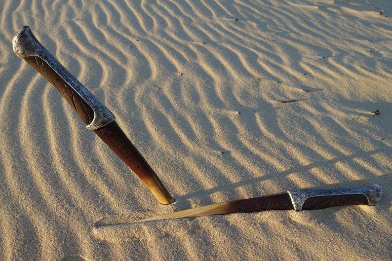 May include: Two wooden-handled knives with silver accents are lying on a sandy surface. The knives are partially buried in the sand.