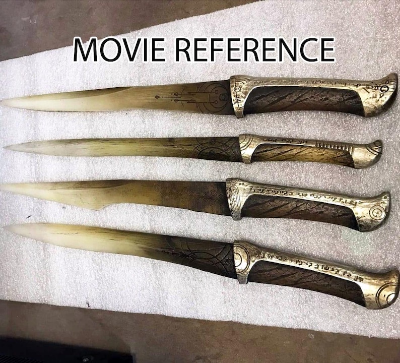 May include: Four ornate, silver-colored daggers with wooden handles. The daggers have intricate designs and patterns etched into the blades and handles. The handles are a dark brown wood with a smooth finish. The daggers are arranged in a row, with the tips pointing to the right. The image is labeled "MOVIE REFERENCE".