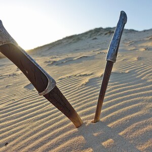 May include: Two silver-colored knives with ornate handles are partially buried in the sand. The knives are pointed towards each other, forming a V-shape.