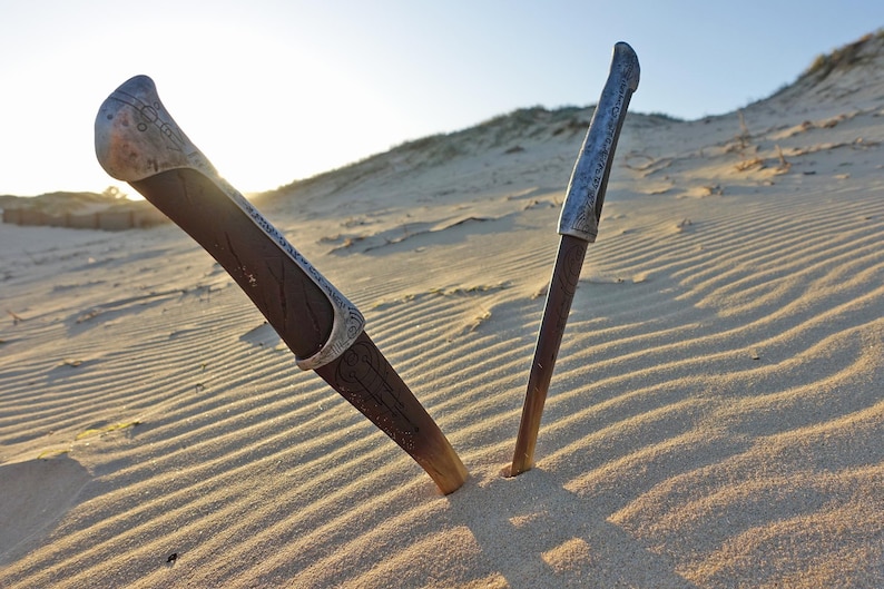 May include: Two silver-colored knives with ornate handles are partially buried in the sand. The knives are pointed towards each other, forming a V-shape.