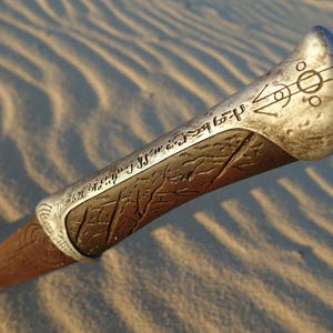 May include: A close-up of a silver and brown metal staff with intricate carvings and a symbol at the top. The staff is lying on a sandy surface.